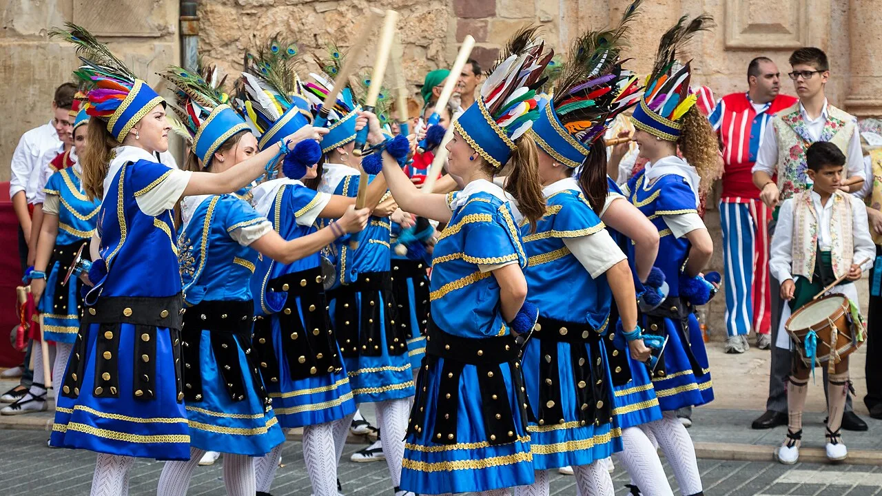 Jóvenes danzantes con traje azul y dorado y altos tocados de plumas entrechocando palos de madera en una calle junto a una iglesia de piedra, con un tamborilero detrás