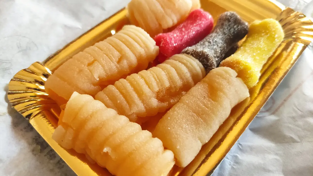 Huesos de santo on a gold cardboard tray: pale ridged marzipan tubes and three sugar-coated ones in red, brown and yellow