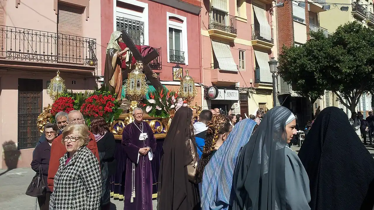 A Semana Santa Marinera procession moving through a street of low painted houses in El Cabanyal, Valencia, with a flower-covered paso carried past the crowd