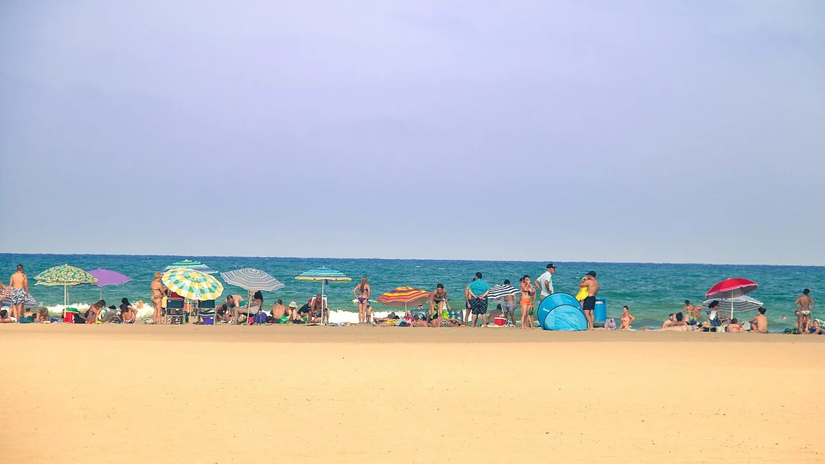 Bañistas y sombrillas sobre la amplia arena de la playa de la Malvarrosa en Valencia, con el mar en calma al fondo