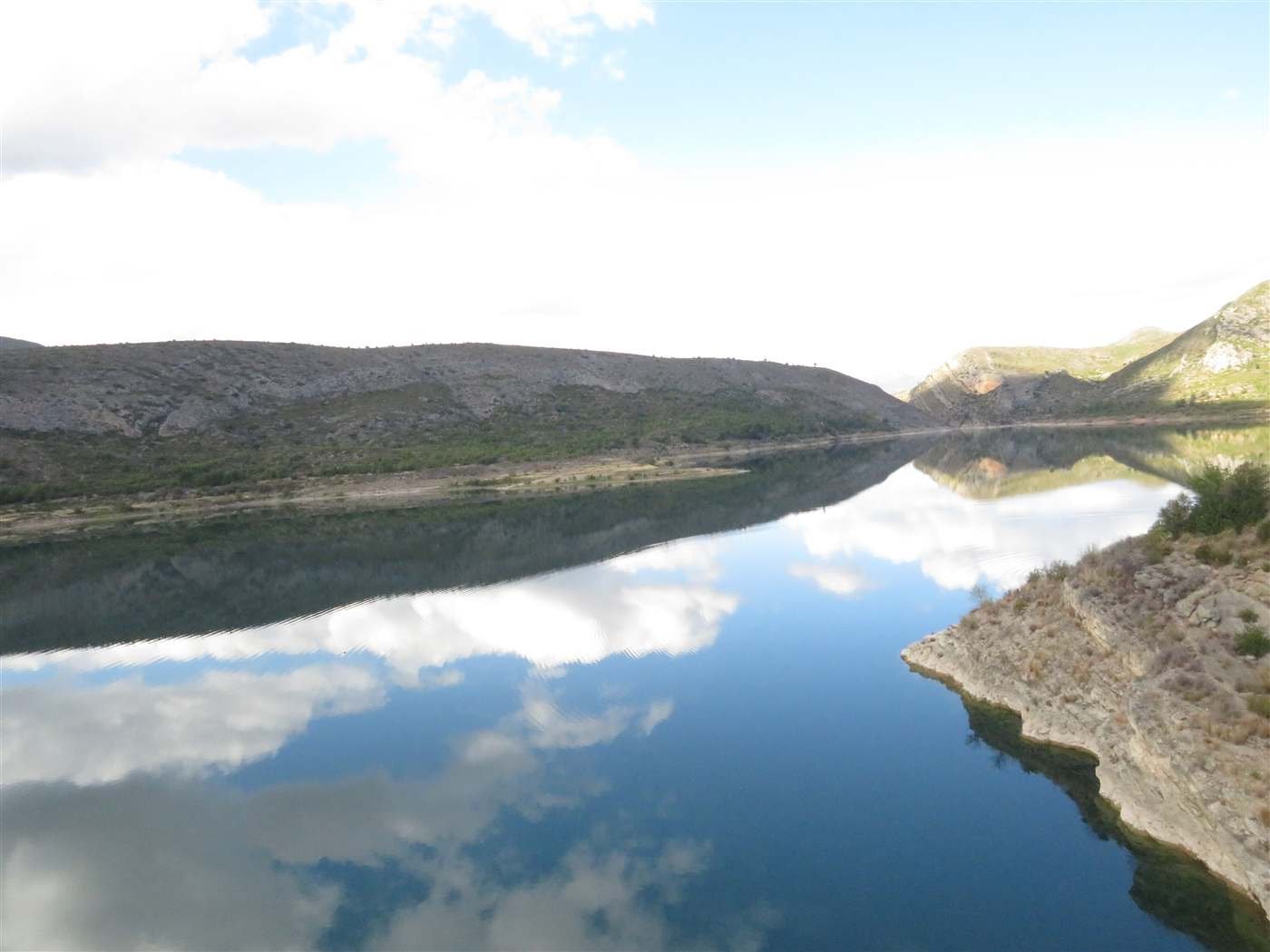 The still, mirror-calm water of the Loriguilla reservoir near Chulilla, reflecting the surrounding hills and clouds.