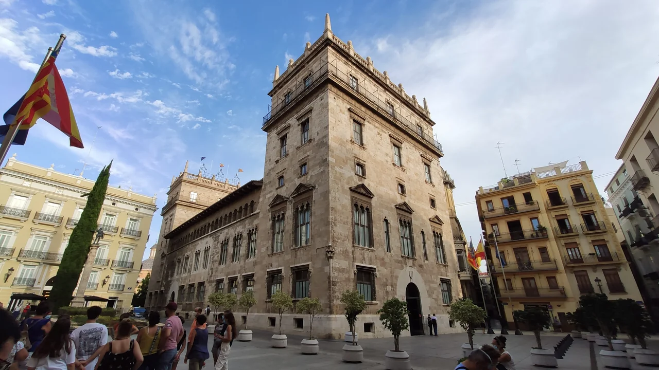 The Gothic stone tower of the Palau de la Generalitat in Valencia under a blue sky, with flags flying at the corner of the square and visitors walking past