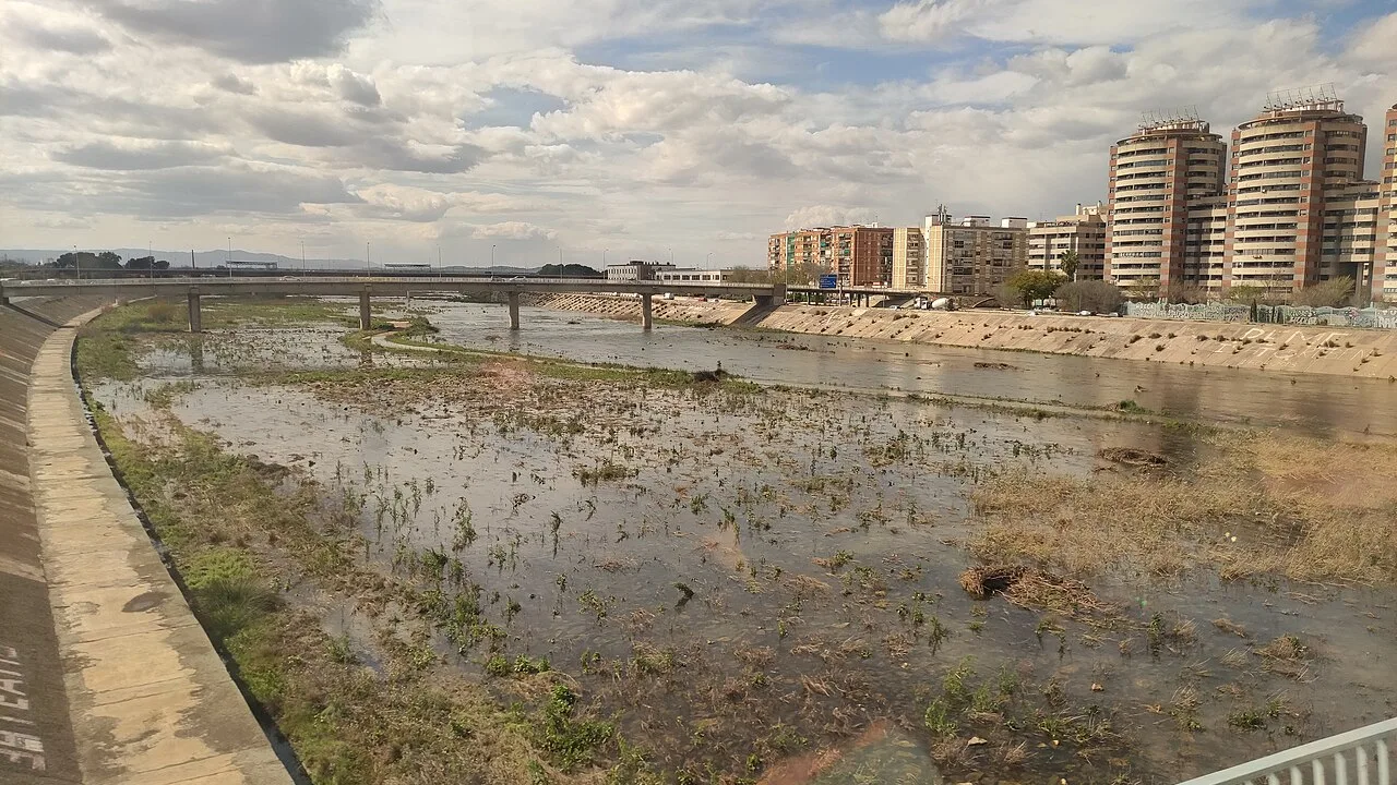 Un large canal artificiel au sud d'une ville espagnole, eau peu profonde et végétation dans son lit, un pont routier le traversant et des immeubles sur la rive opposée