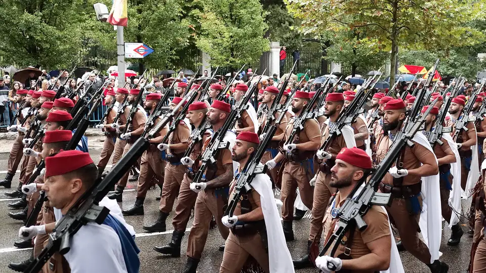 Soldiers of the Regulares in red caps and white capes marching with rifles on a wet Madrid avenue, crowds with umbrellas behind them