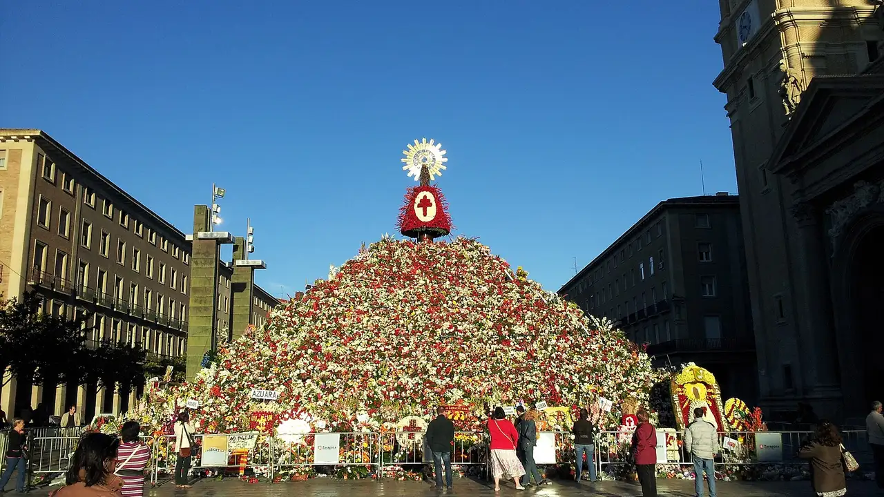 A tall pyramid of red and white flowers topped by a small image of the Virgin in the Plaza del Pilar, Zaragoza, with the basilica tower on the right