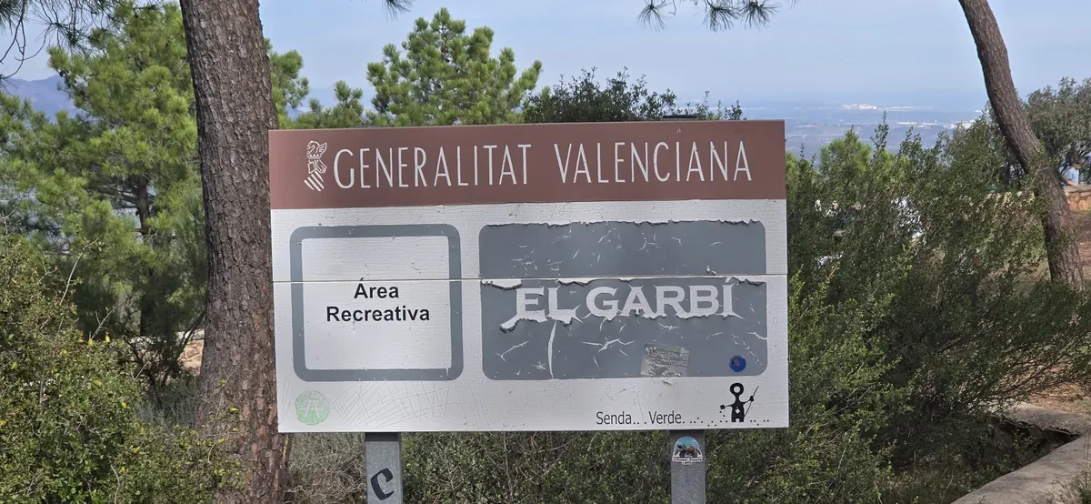 Generalitat Valenciana sign for the Àrea Recreativa El Garbí among pines, with the coastal plain and the sea behind