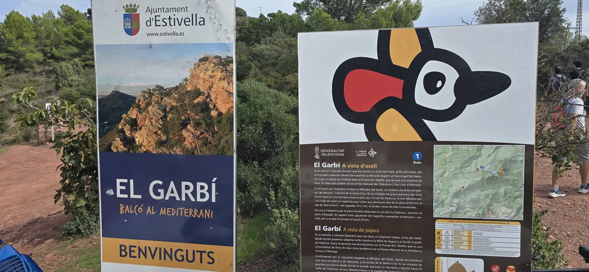Two trailhead panels near the top of the Garbí: the Ajuntament d'Estivella welcome board reading El Garbí, balcó al Mediterrani, and the Generalitat's Serra Calderona route panel with a map