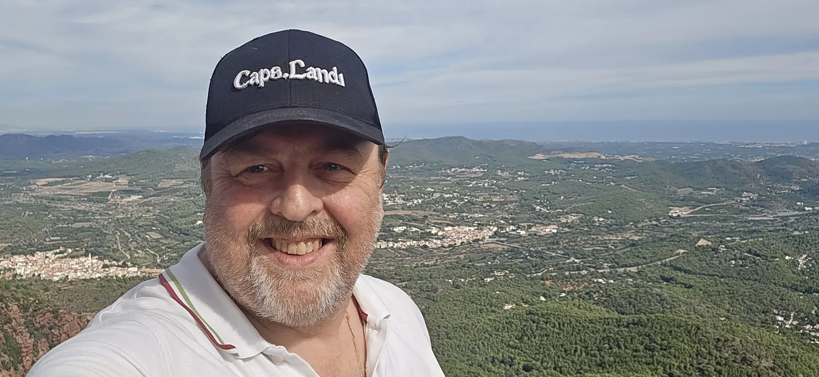 Michael Bastin met pet lachend op de top van de Garbí, met beboste heuvels, dorpen, de kustvlakte en de zee achter hem