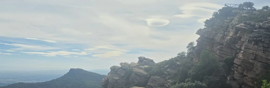 De Mirador del Garbí met reling boven op gelaagde rode zandsteenkliffen, met de Mola de Segart met vlakke top en de kustvlakte onder lensvormige wolken