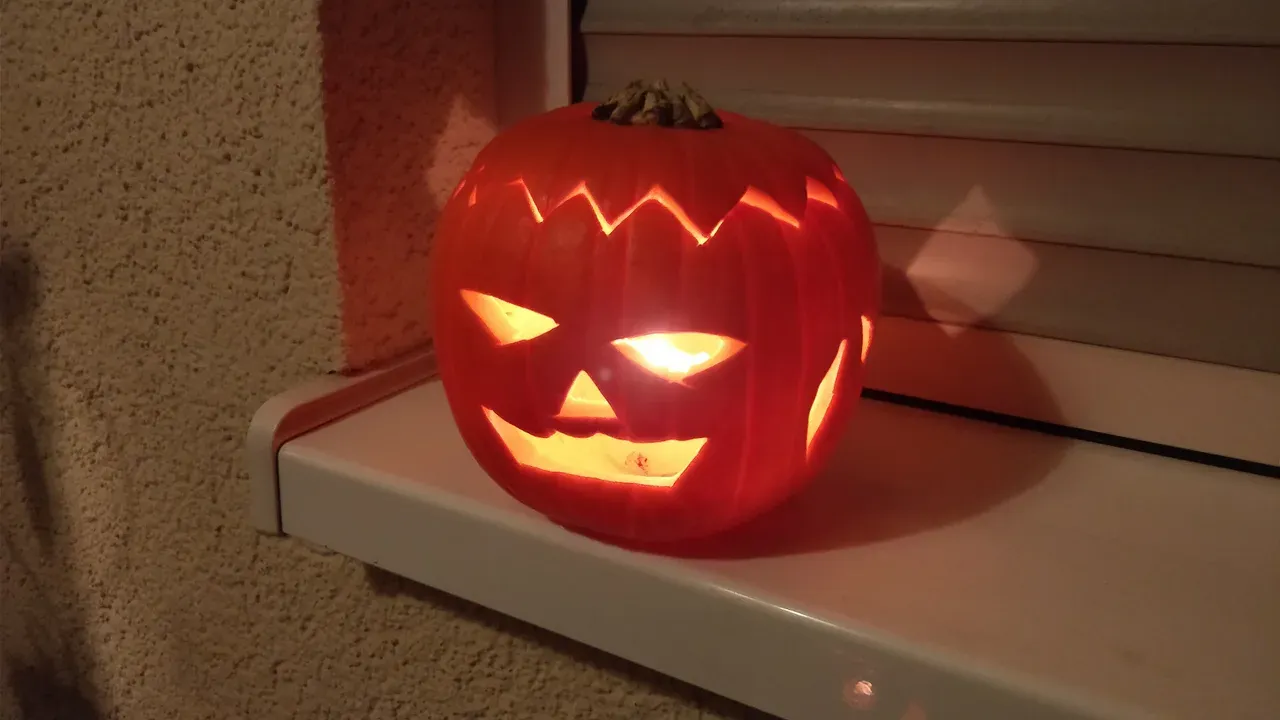 Una calabaza tallada iluminada con una vela, sobre el alféizar blanco de una ventana delante de una persiana, de noche