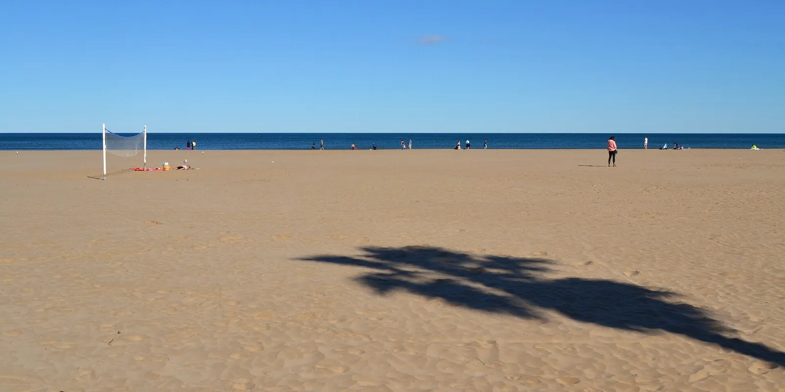 Bijna leeg strand van Patacona in Alboraya met een volleybalnet, een paar wandelaars en de schaduw van een palmboom op het zand