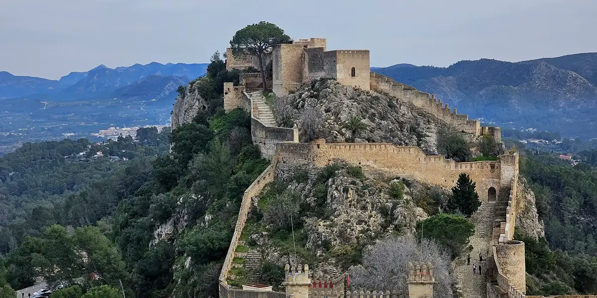 Het Castell de Xàtiva langs de rotsige bergkam, met muren en torens boven beboste hellingen en bergen op de achtergrond