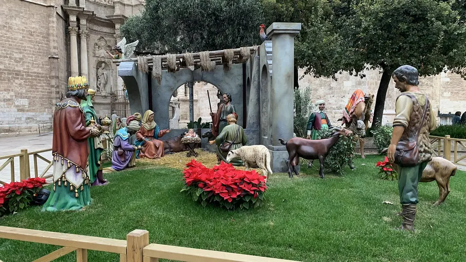 Une crèche grandeur nature en plein air sur une pelouse, avec les Rois mages, la Sainte Famille, des bergers et des animaux, des poinsettias rouges au premier plan et la façade de pierre de la cathédrale de Valencia derrière
