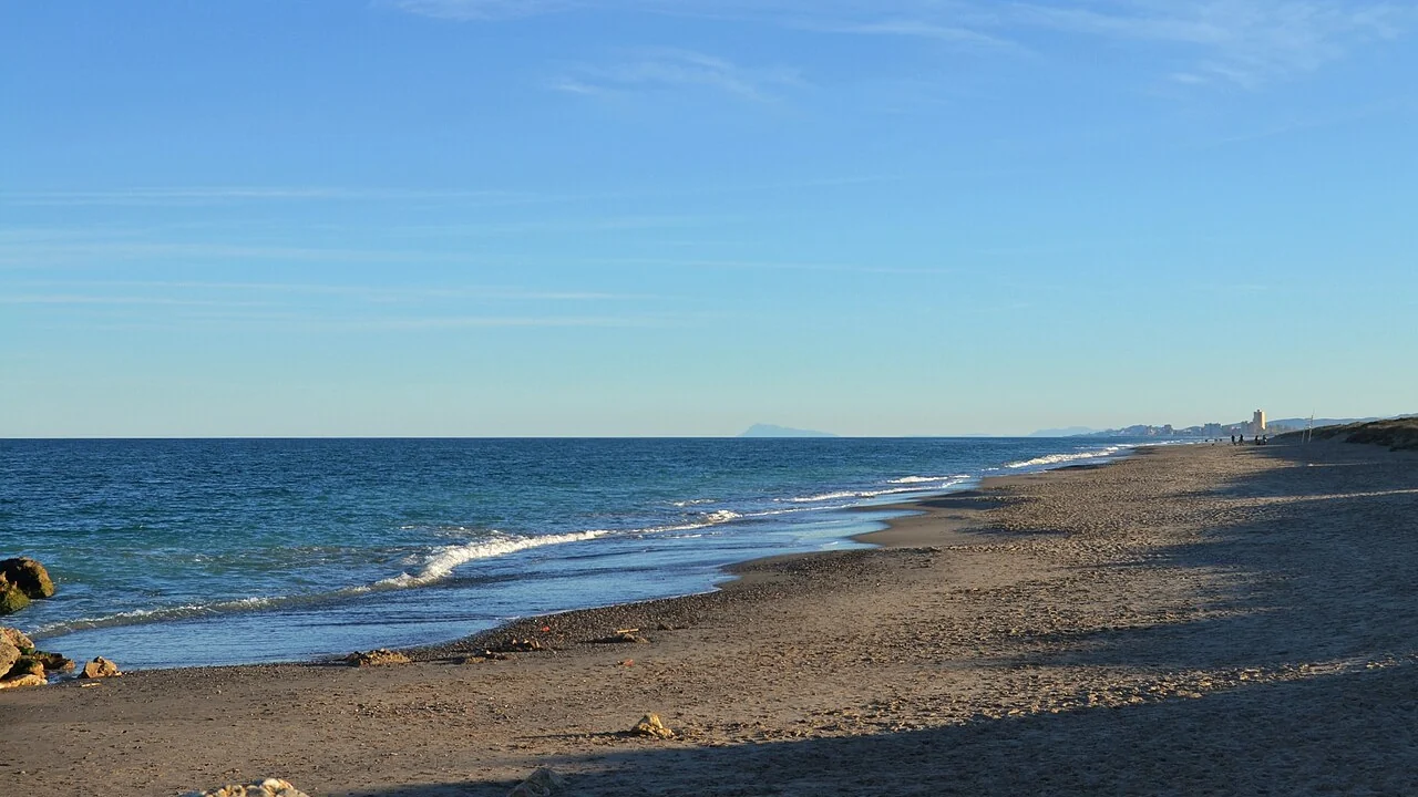 The wide, undeveloped dune beach of the Devesa del Saler in low sun, with rocks in the foreground and no buildings along the shore.