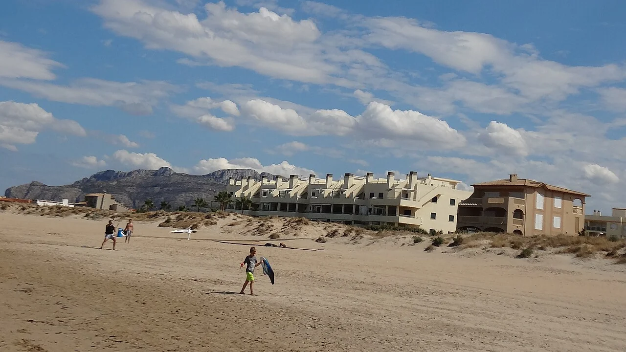A kitesurfer carrying a board across the wide dune-backed sand at Oliva, with low apartment buildings and the Montgo massif behind.