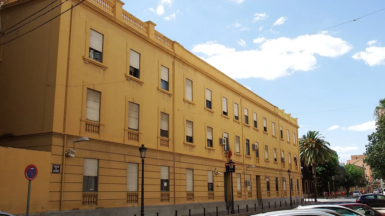 The long yellow facade of a Spanish national police station on a city street, shuttered windows and parked cars along the kerb under a bright sky