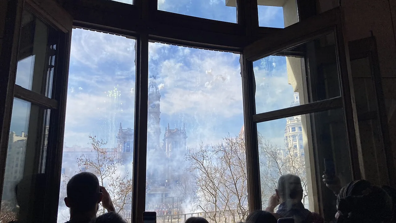 La mascletà vista a través de una ventana sobre la plaza del Ayuntamiento de Valencia, la plaza llena de humo y la torre del consistorio detrás