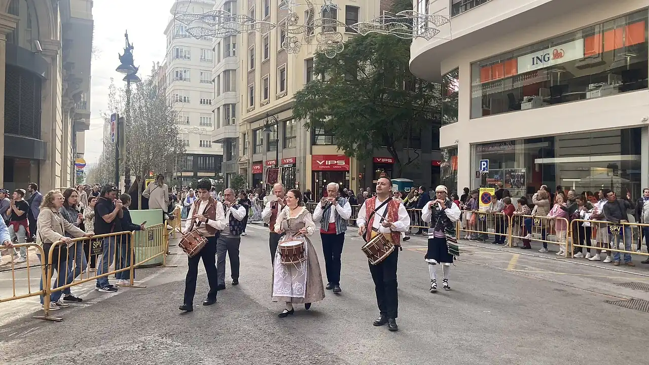 Musicians in traditional Valencian dress playing drums and wind instruments in the Fallas flower offering procession, with spectators behind barriers