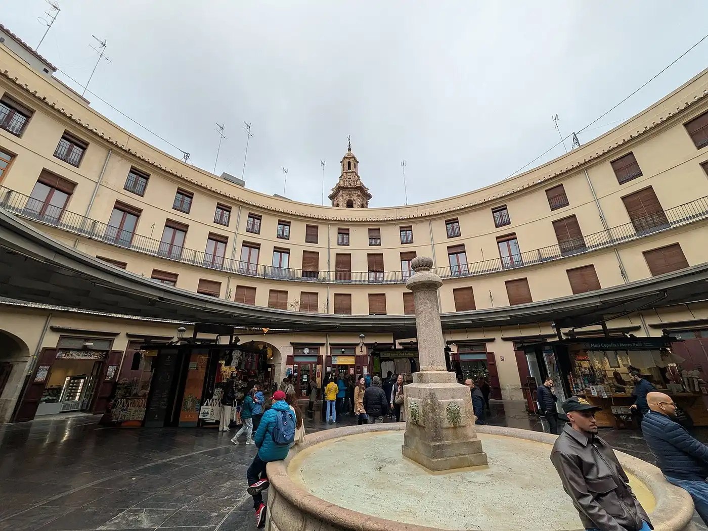 The circular courtyard of the Plaza Redonda in Valencia, ringed by balconied buildings and ground-floor stalls, with the stone fountain in the centre and the Santa Caterina bell tower behind