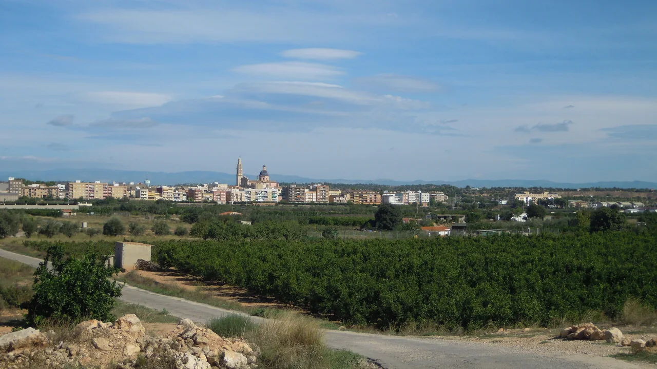 La ville de Cheste vue au-delà de champs verts et d'un chemin, avec un clocher et un dôme qui dépassent des immeubles