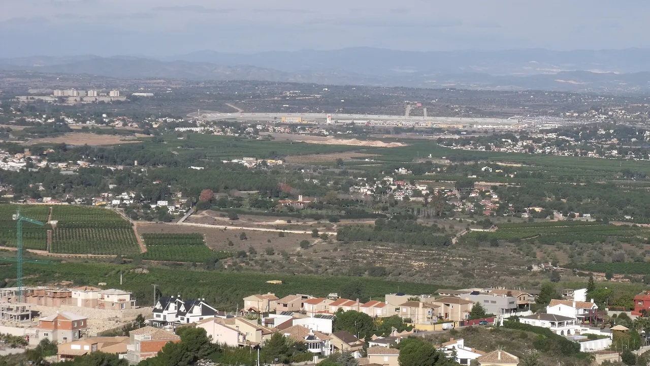 Vue large sur des villas, des vergers et des champs jusqu'aux longues tribunes grises du Circuit Ricardo Tormo, avec des montagnes à l'horizon