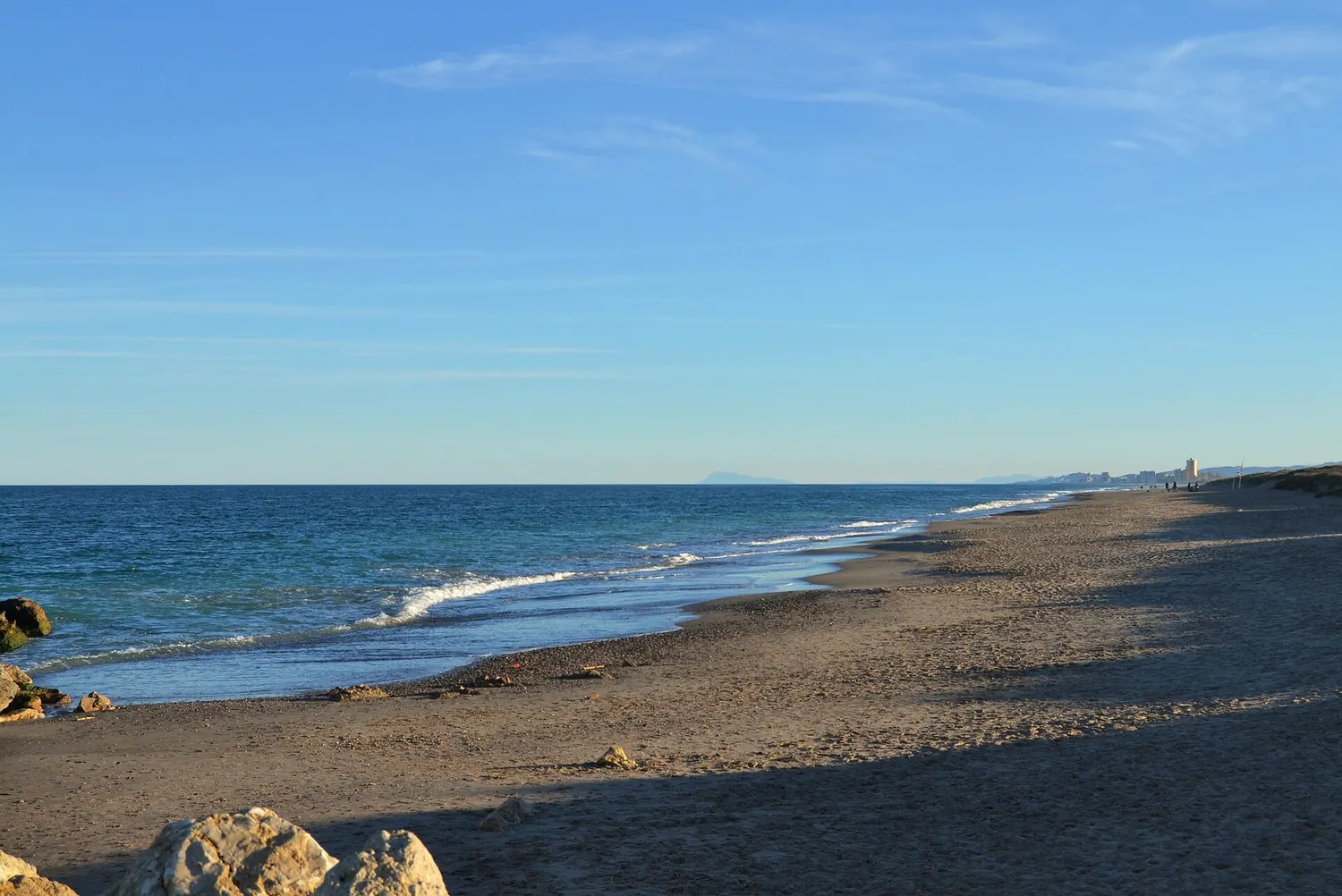 Une longue plage de sable déserte avec de petites vagues et quelques rochers au premier plan, bordée de dunes, à la Devesa del Saler au sud de Valencia