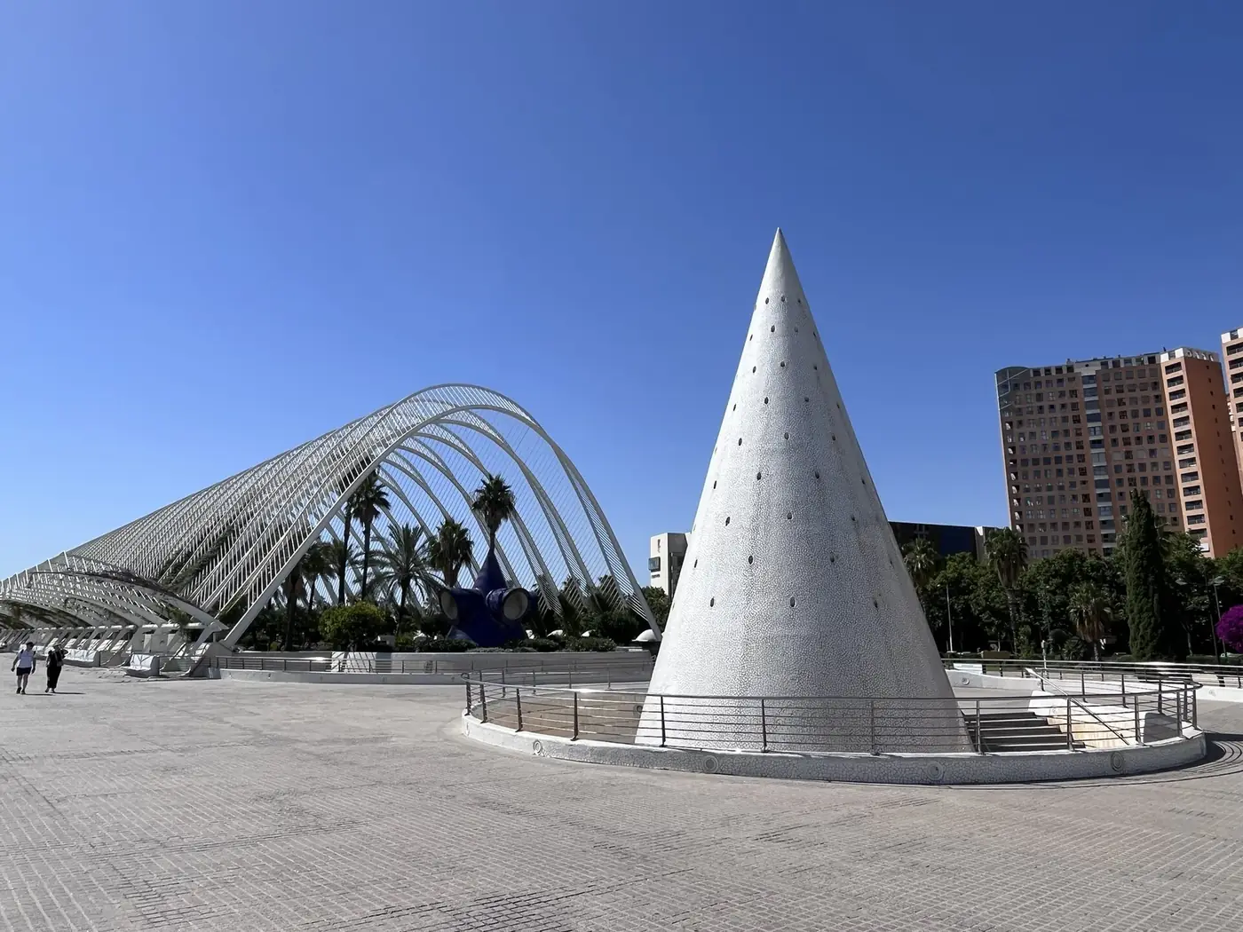 La structure blanche en arcs de L'Umbracle à côté d'un cône blanc sur une place dallée de la Cité des Arts et des Sciences, sous un ciel bleu dégagé