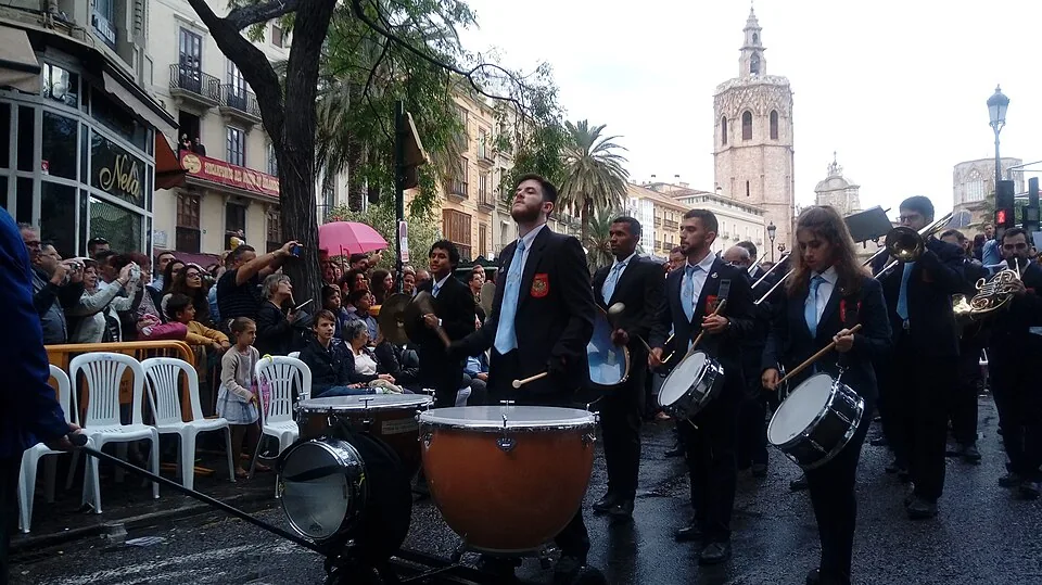 Muziekkorps met trommels, pauken, een trombone en een hoorn in een straatparade, met de klokkentoren Micalet achter toeschouwers op witte stoelen