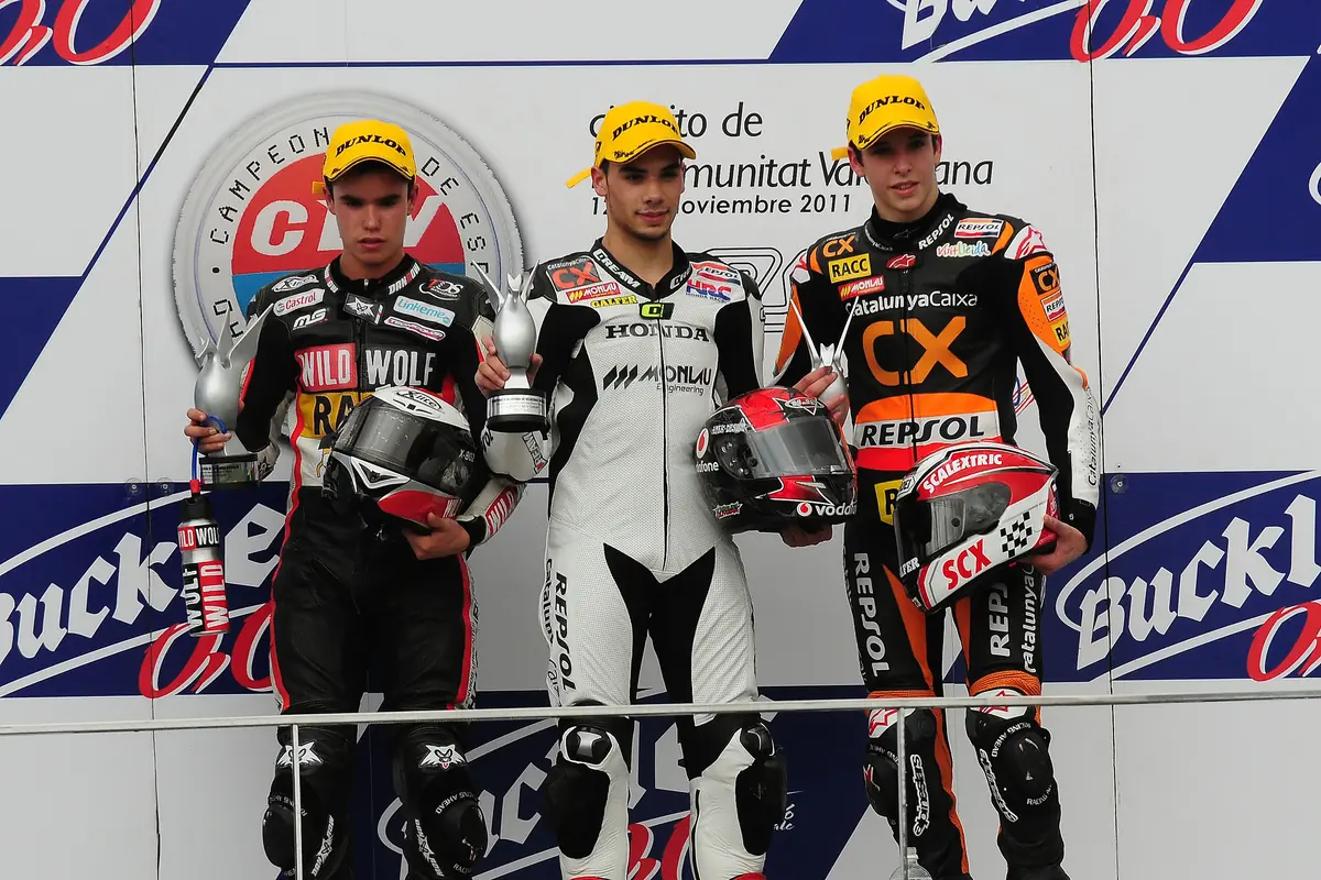 Drie motorcoureurs in leren pakken op het podium van het Circuit Ricardo Tormo in Cheste, met sponsorborden achter hen