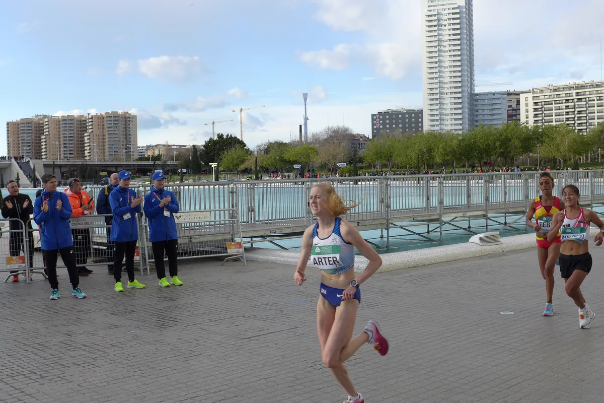 Una corredora con la camiseta de Gran Bretaña encabeza un grupo de atletas por el recorrido del medio maratón de Valencia junto a un canal, con torres de viviendas detrás