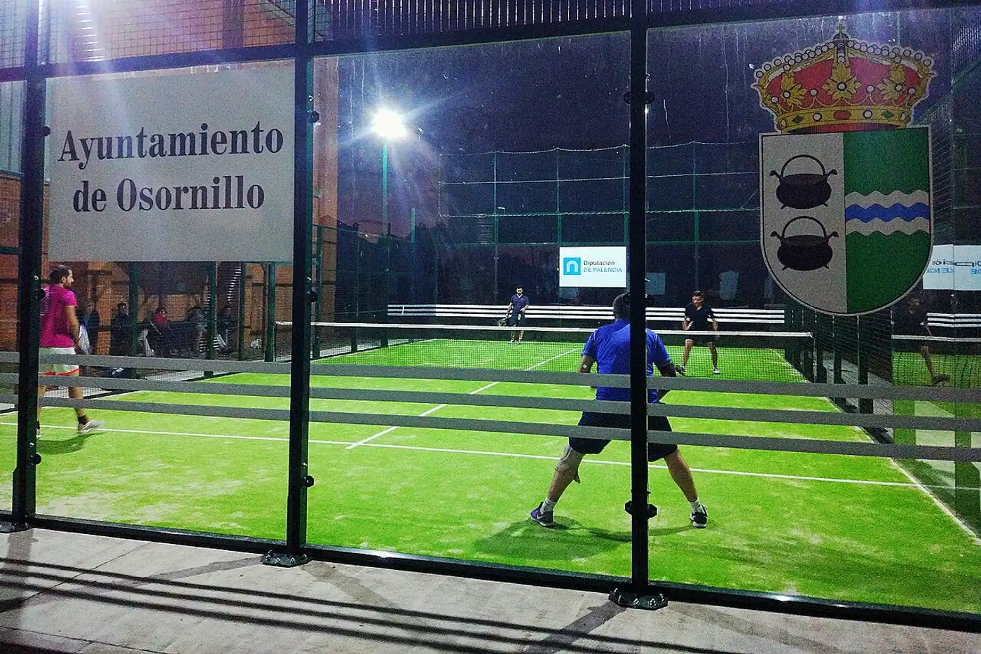 Una pista de pádel exterior en España de noche, con dos jugadores en pleno partido tras los cristales y la malla.