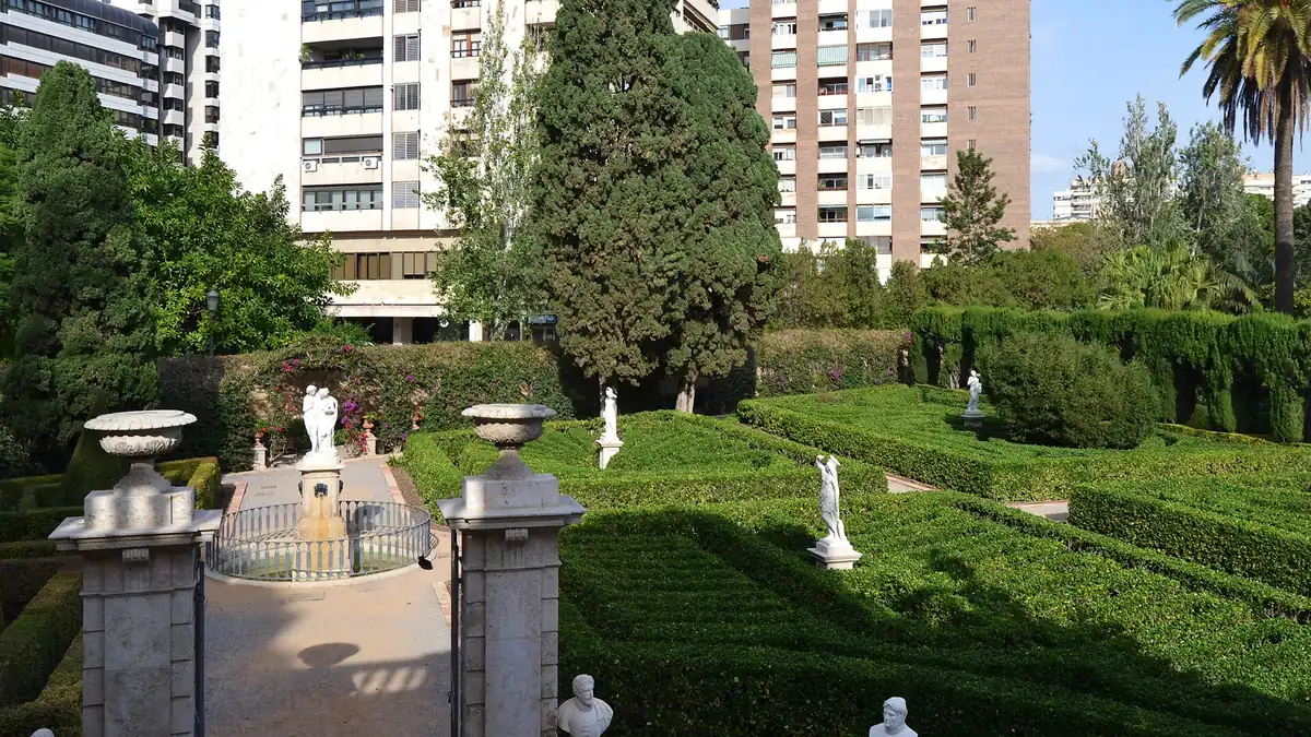 Geschoren buxushagen, marmeren beelden en een fontein in de Jardines de Monforte, met moderne woontorens achter de tuinmuur