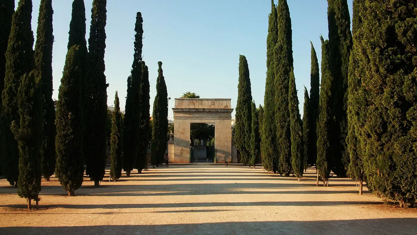Een laan met hoge cipressen die naar een neoklassieke stenen poort leidt in de Jardines del Real, Valencia