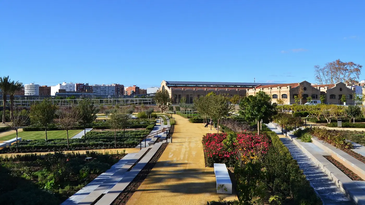 Grindpaden, kruidenbedden en bloeiende struiken in Parc Central Valencia, met de gerestaureerde bakstenen spoorwegwerkplaatsen erachter