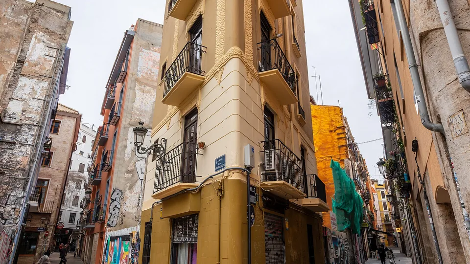 Een smal crème en okergeel hoekgebouw met smeedijzeren balkons waar twee geplaveide steegjes in de oude stad van Valencia samenkomen, met een voorbijgangster met een boodschappentas