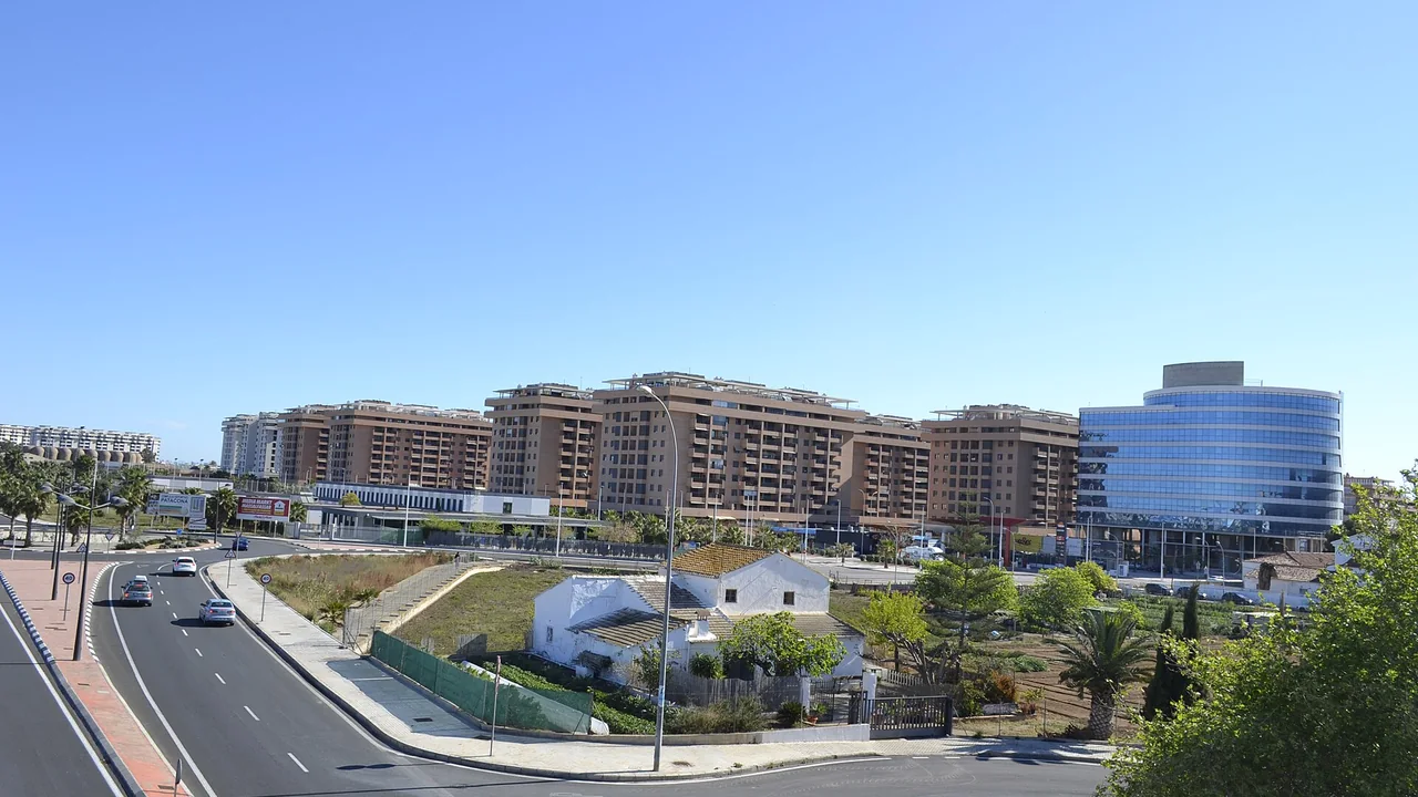 Een rij nieuwe bruine appartementsgebouwen en een glazen kantoorgebouw achter een oude witte boerderij en een brede weg aan de rand van Valencia