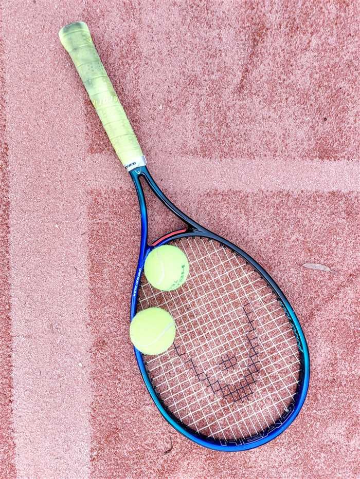 A tennis racket and two balls resting on the red clay of a court, with the white sideline visible