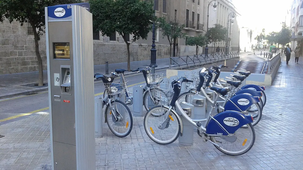 Une station Valenbisi avec une rangée de vélos bleus à panier et une borne de paiement dans une rue de Valencia