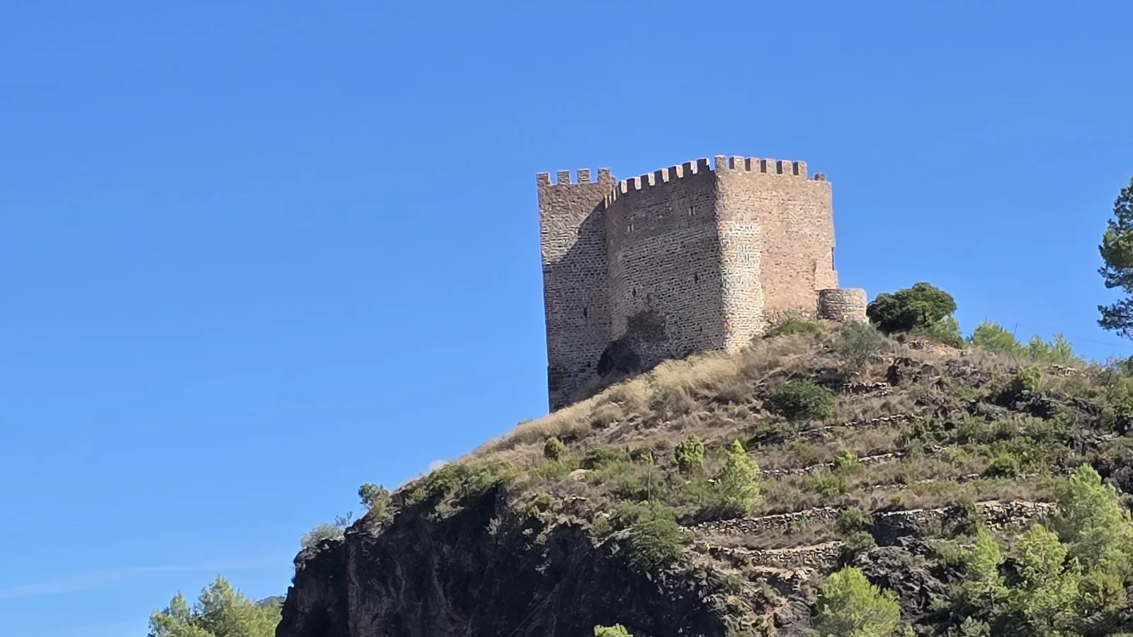 Le donjon crénelé en pierre du château de Gaibiel sur un éperon rocheux couvert de pins, sous un ciel bleu