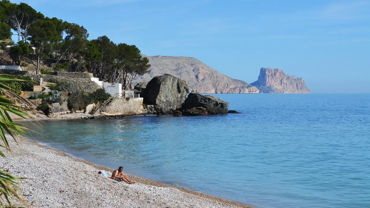 Une plage de galets à Cap Negret, à Altea, avec des pins, un gros rocher et le Penyal d'Ifac qui se dresse dans la mer au loin