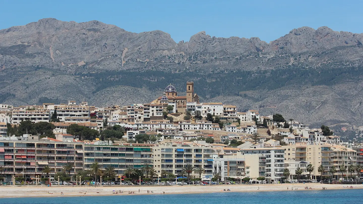 Altea vue depuis le port : immeubles et plage en front de mer, la vieille ville blanche sur la colline avec le dôme bleu de l'église, et des montagnes nues derrière