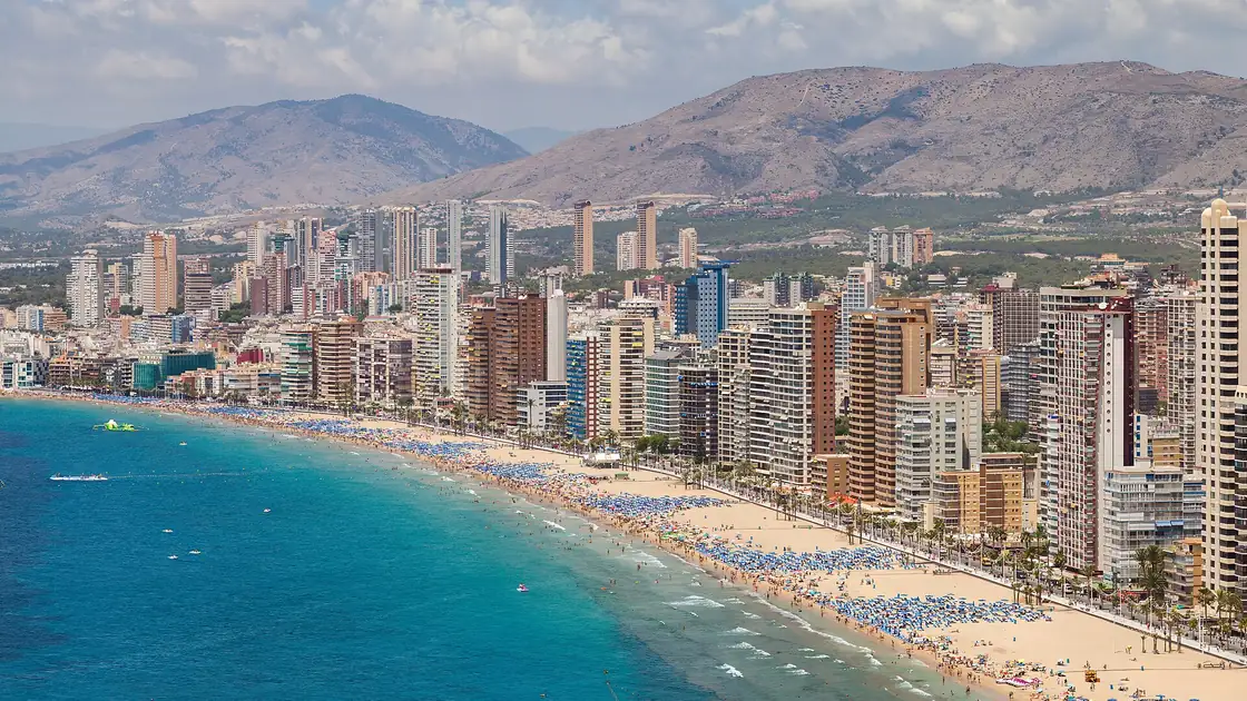 La plage de Levante à Benidorm : une longue courbe de sable doré avec des parasols, une promenade bordée de palmiers et des tours, avec des collines derrière