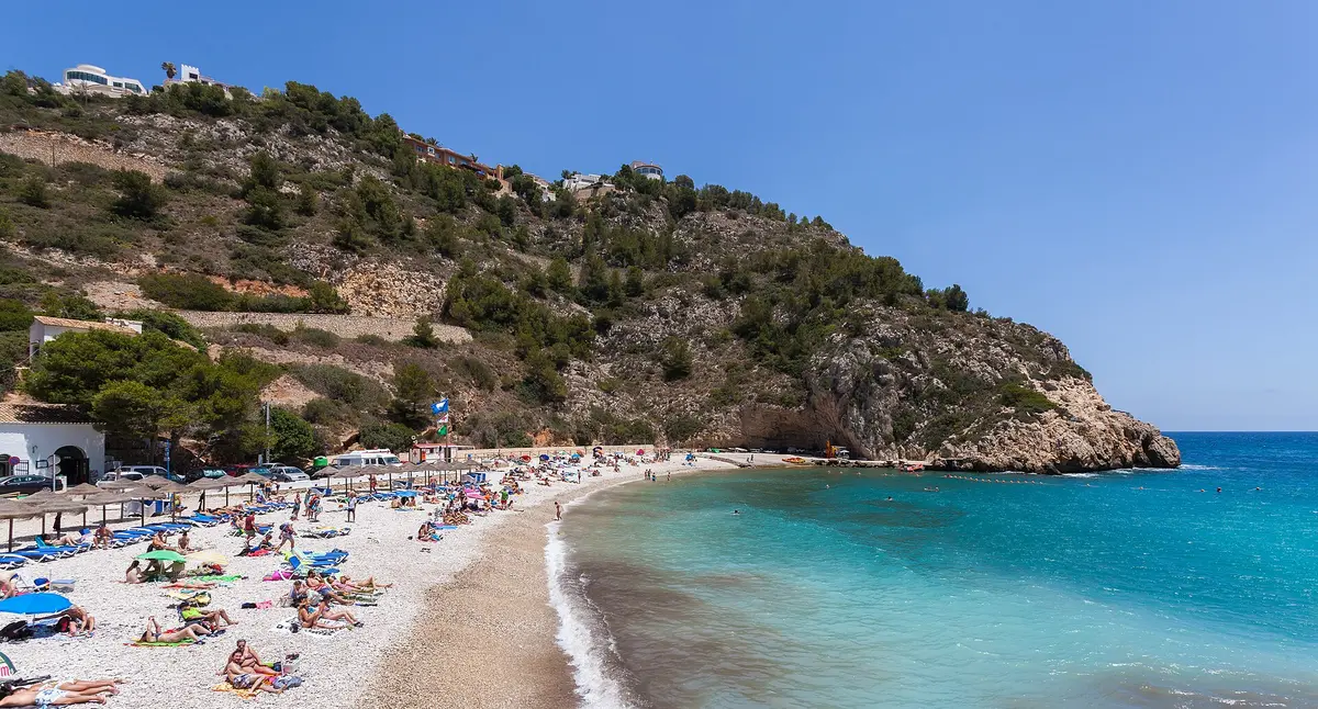 La Cala de la Granadella à Xàbia : plage de galets avec transats sous un promontoire couvert de pins et une eau turquoise