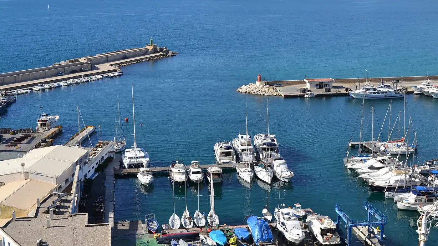 Le port de Calpe vu d'en haut, avec des bateaux de pêche et des voiliers amarrés aux pontons, des digues aux feux rouge et vert et la côte vers Altea au fond