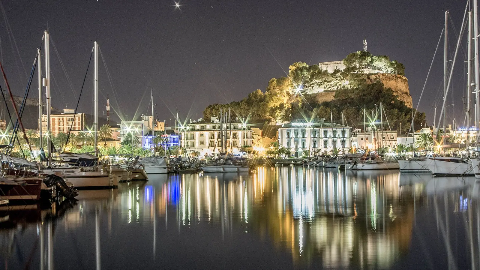 Dénia castle lit up at night on its hill above the town, with sailing boats in the marina reflected in the water