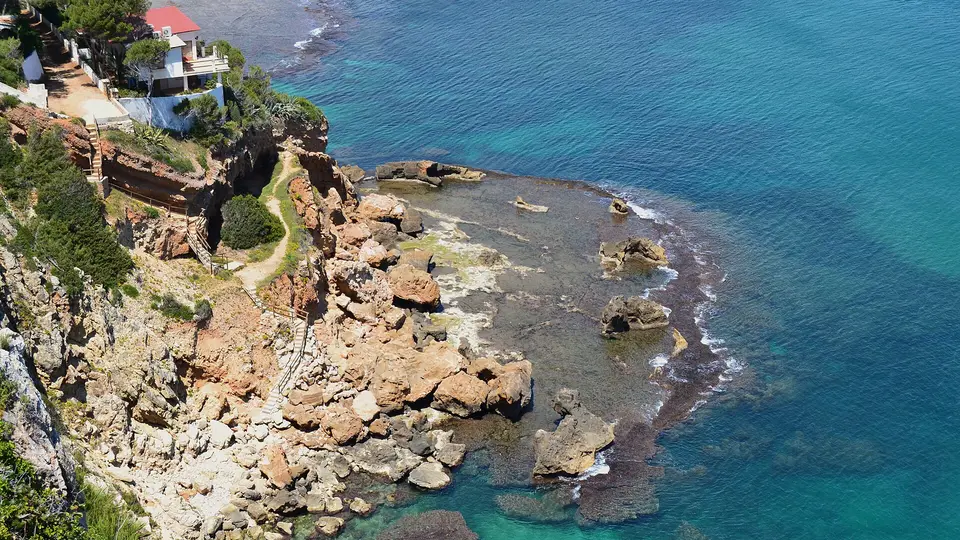 The rocky coast of Les Rotes in Dénia seen from above, with a house among pines, a path down the cliff and clear turquoise water