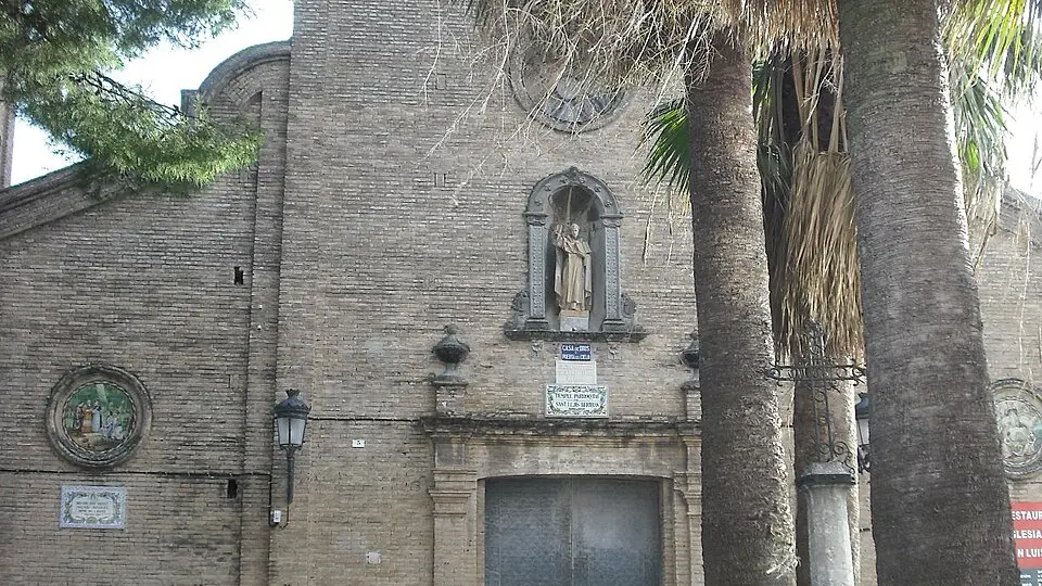 La façade en brique de l'église de Sant Lluís Bertran, avec une statue du saint dans une niche au-dessus de la porte et des palmiers à côté, à La Fonteta de Sant Lluís, Valencia