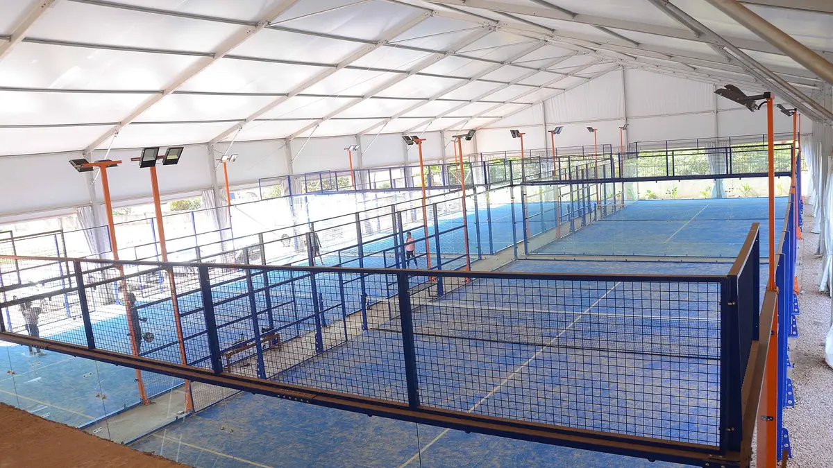 Rangée de terrains de padel bleus avec murs du fond vitrés et côtés grillagés sous une toiture tendue blanche