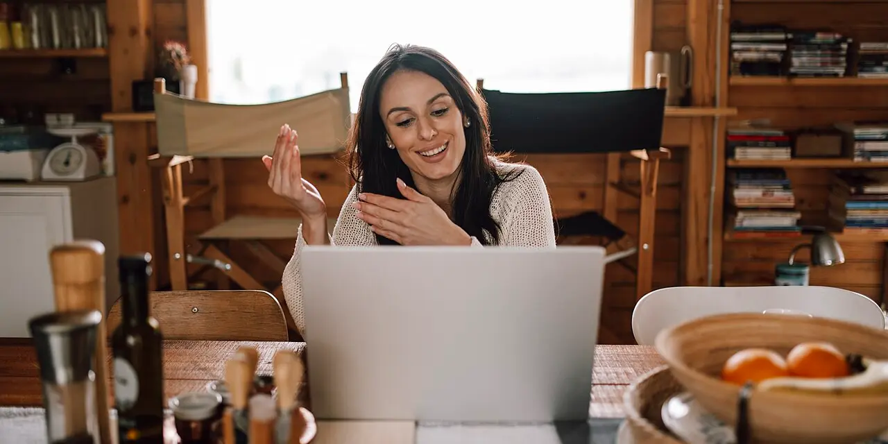 Een lachende vrouw in een videogesprek op een laptop aan een houten keukentafel