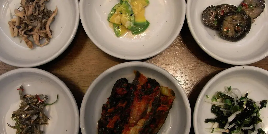 Seis cuencos blancos de banchan coreano sobre una mesa de madera, con kimchi y verduras aliñadas